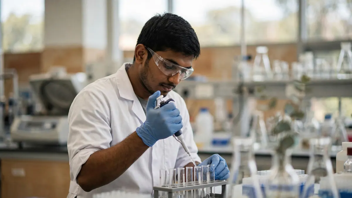 A lab technician in safety glasses and blue gloves pipettes a sample into a rack of test tubes