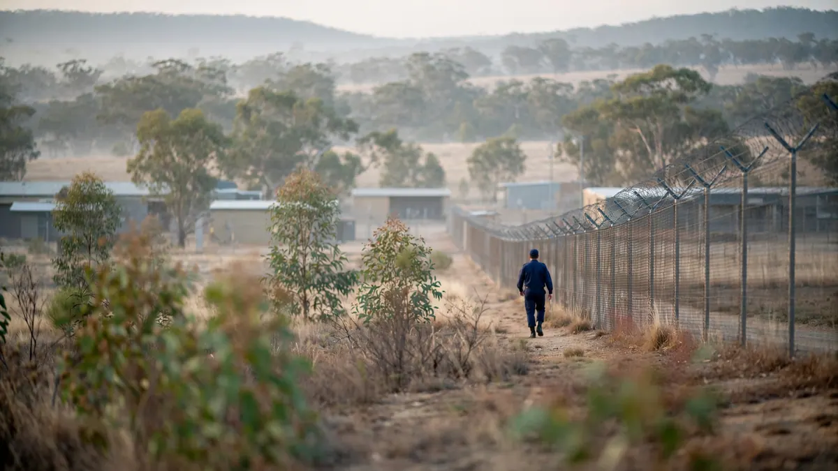 An officer in a navy uniform walks the wire perimeter fence of a correctional centre in morning mist